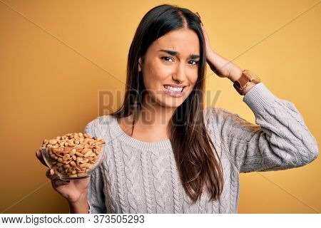 Young beautiful brunette woman holding bowl with peanuts over isolated yellow background stressed with hand on head, shocked with shame and surprise face, angry and frustrated. Fear and upset.