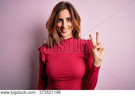 Young beautiful brunette woman wearing casual sweater stnding over pink background smiling with happy face winking at the camera doing victory sign. Number two.