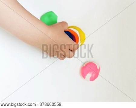 Toddler Sculpts From Colored Plasticine On A White Table. The Hand Of A Small Child Squeezes Pieces 