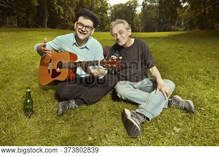 Two Men On City Park Summer Meadow Enjoying Day With Guitar