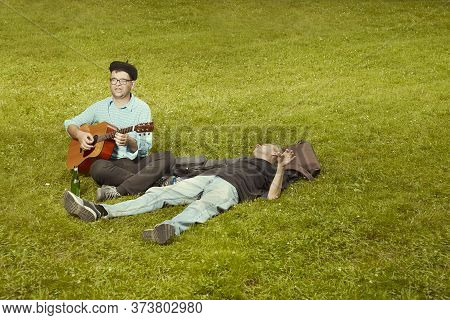 Two Men On City Park Summer Meadow Enjoying Day With Guitar