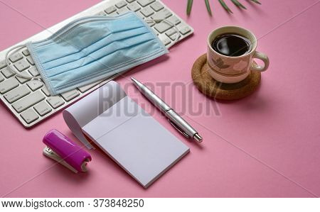 Medical Face Mask On Top Of Keyboard With Coffee And Stationery On Pink Colofr Office Desk.