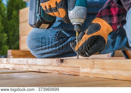Male Carpenter Using Cordless Drill To Drive Long Screw Through Wooden Planks.