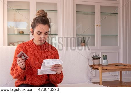 Focused Woman Examining Drug Package Leaflet For Prescribed Medicine While Sitting At Home Holding B