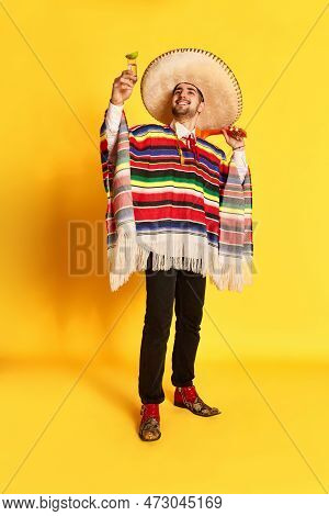 Cheers. Young Smiling Man In Colorful Festive Clothes, Poncho And Sombrero Posing With Tequila And L
