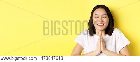 Close Up Of Hopeful Asian Girl In White T-shirt, Holding Hands In Pray, Namste Gesture And Smiling, 