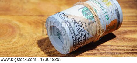 A Roll Of Dollars On A Wooden Textured Table In The Dark Illuminated By A Ray Of Light.