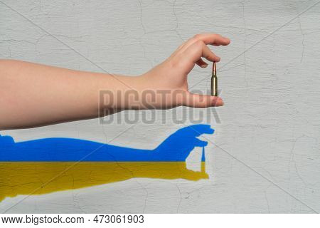 Rifle Cartridge In Hand On The Background Of The Wall. A Shadow Of Yellow-blue Color (national Symbo