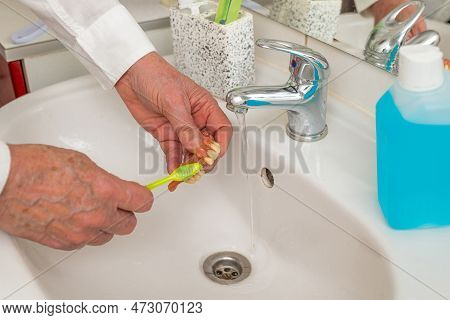 Hands Of A Man Cleaning Dentures With A Toothbrush In The Sink Under The Tap. Close Up.