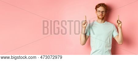 Confident And Pleased Young Man With Red Hair, Wearing Glasses And T-shirt, Pointing Fingers Up And 