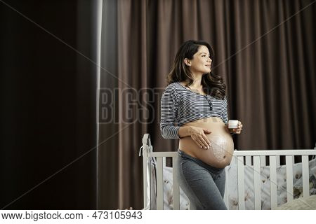 A Happy Pregnant Woman Is Putting On Anti-stretchmarks Lotion In A Bedroom While Standing Next To A 