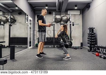 A Young Male Personal Trainer Helps A Woman In An Electric Muscle Suit To Stimulate The Muscles Whil