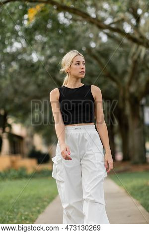 Young Blonde Woman In White Cargo Pants And Black Tank Top Walking On Green Street In Miami.