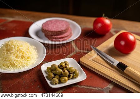 Different Ingredients For Making Pizza - Grated Cheese, Olives, Salami, Tomatoes On The Kitchen Tabl