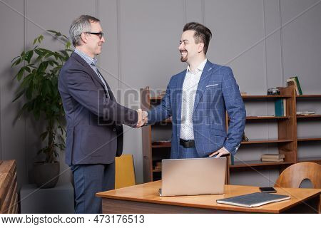 Two Happy Cherrful Businessman In Blue Suits Smiling And Making Handshake In Office By The Laptop, B