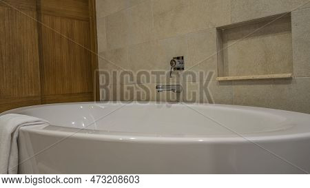 Large Acrylic Oval Bathtub, Close-up. A White Towel Is Thrown Over The Edge. On The Beige Tiled Wall
