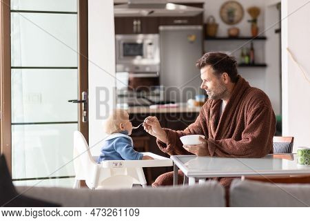Father Wearing Bathrope Spoon Feeding Hir Infant Baby Boy Child Sitting In High Chair At The Dining 