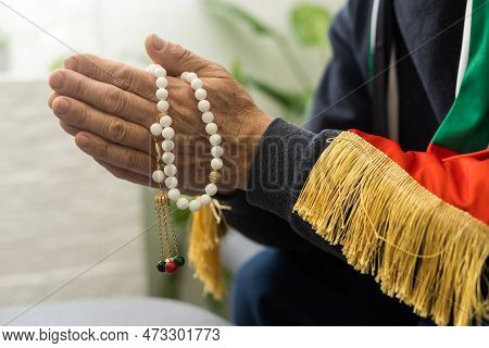 An Elderly Man Prays With A Rosary And The Flag Of The United Arab Emirates