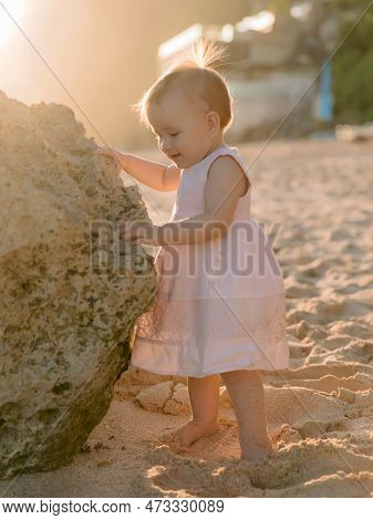 Portrait Of Charming Baby Girl In Pink Dress On Sandy Beach With Sunset Tones.