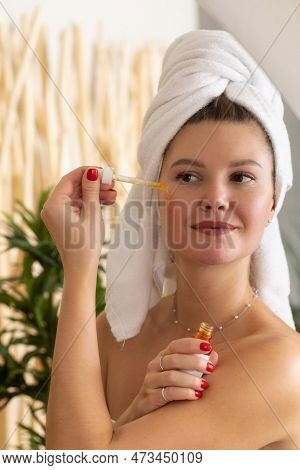 An Attractive Woman In Front Of A Mirror Applies A Serum To The Skin Of The Face In The Bathroom.
