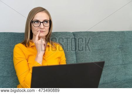 Puzzled Woman In An Orange Shirt And Glasses Is Sitting On The Sofa With A Laptop, Thinking Intently