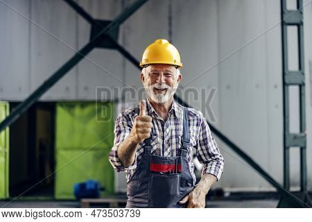 Good Business In The Factory. A Satiated Smiling Senior Worker With A Helmet On His Head Is Standing
