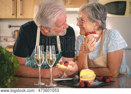 Happy Senior Couple In Home Kitchen Looking In The Eyes Eating A Handmade Plumcake With Berries