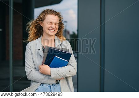 Portrait Of Woman Student Standing Outdside The University Campus Wearing Jacket, Holding Books And 