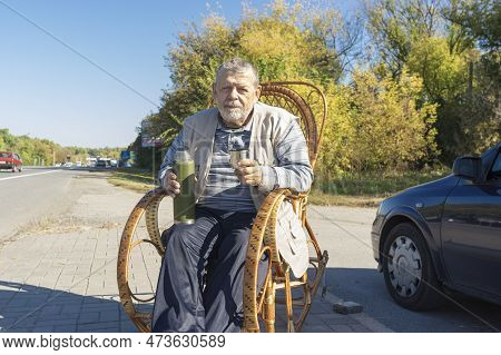 Portrait Of Ukrainian Senior Man Sitting In Rrocking Chair On Roadside And Holding Vacuum Bottle Get