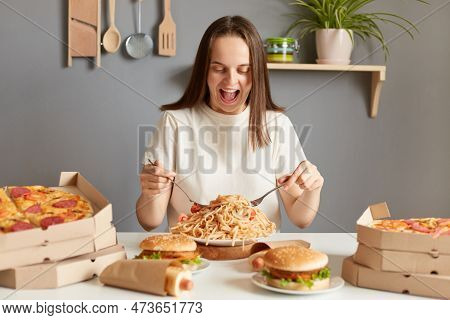 Portrait If Amazed Excited Extremely Hungry Woman Wearing White T-shirt Sitting At Table In Kitchen,