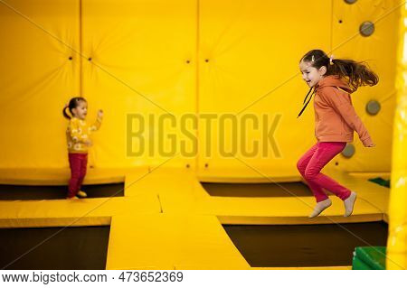 Siblings Jumping On Trampoline At Yellow Playground Park. Sisters In Motion During Active Entertaime