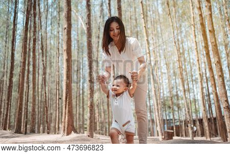 Baby Taking First Steps In Forst On Meadow Grass. Mother Supports Child To Learn Walking Forward, Ba