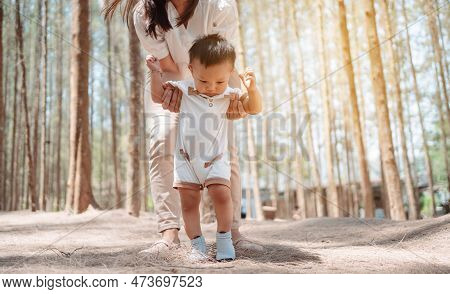 Baby Taking First Steps In Forst On Meadow Grass. Mother Supports Child To Learn Walking Forward, Ba