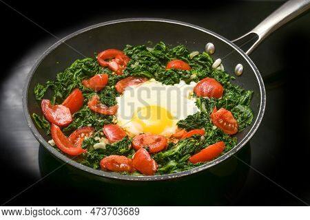 Fried Egg With Spinach And Tomatoes In A Frying Pan On A Black Cooker Top, Preparing A Healthy Meal 