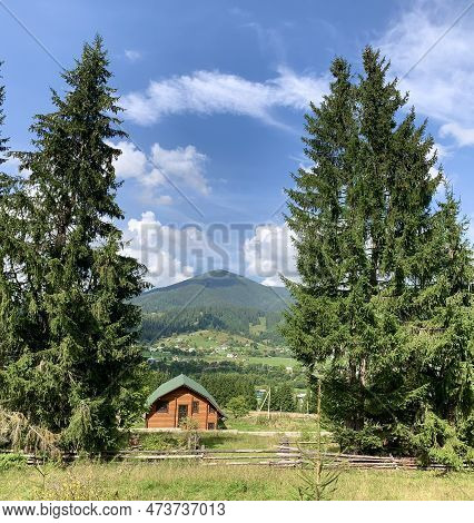 Wooden House Against The Backdrop Of Firs And High Mountains. Tall Coniferous Trees On The Backgroun