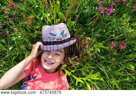 Portrait Of A Child In A Hat With His Face Covered In Summer Lying In The Grass And Wildflowers. Hat