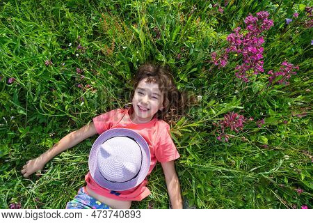 Portrait Of A Child In A Hat With His Face Covered In Summer Lying In The Grass And Wildflowers. Hat