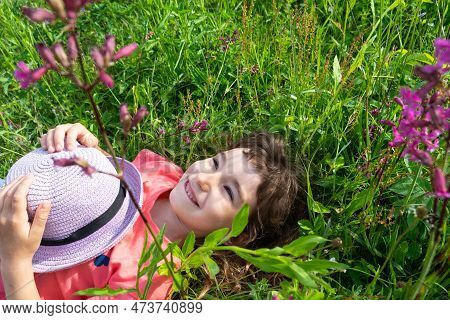 Portrait Of A Child In A Hat With His Face Covered In Summer Lying In The Grass And Wildflowers. Hat