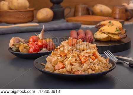 Stewed Cabbage - Bigos In A Black Plate Against The Background Of Pickled Vegetables And Baked Potat