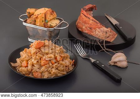 Stewed Cabbage - Bigos In A Black Plate On The Background Of A Piece Of Smoked Meat And Baked Potato