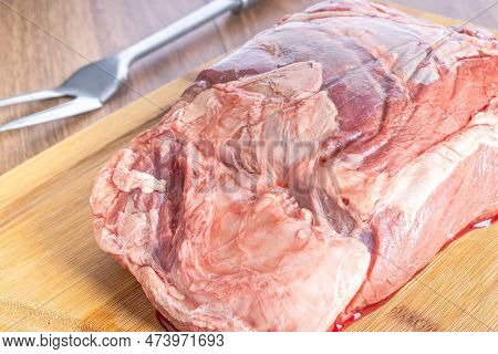 Whole Raw Sirloin Steak On A Wooden Board, On A Wooden Table. With Large Stainless Steel Fork.