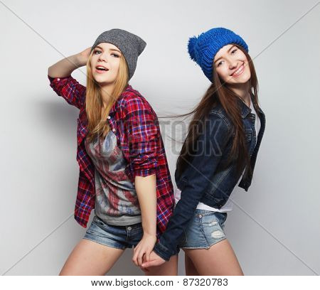 Fashion portrait of two stylish sexy hipster girls best friends, wearing cute swag outfits and hats. Over gray backround.