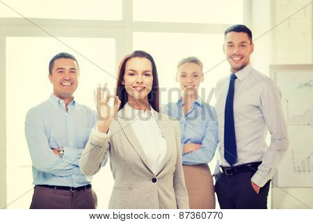 office, business and teamwork concept - friendly young smiling businesswoman with team on back showing ok-sign