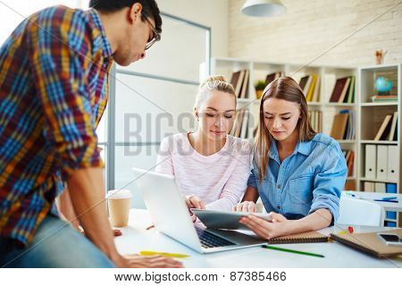 Two cute teenage girls looking through data in touchpad with their groupmate near by