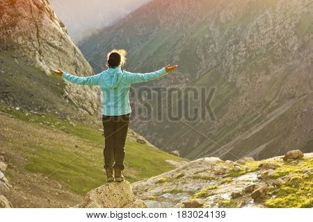 girl in blue jacket standing with hands up above mountain slopes at yellow sunset