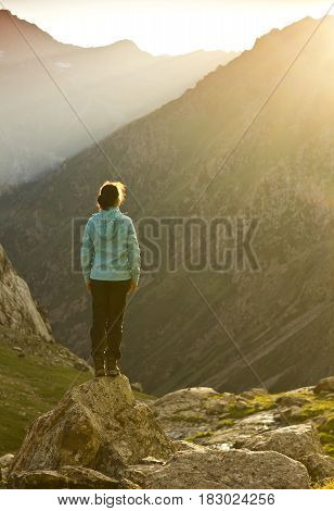 girl in blue jacket standing with hands up above mountain slopes at yellow sunset