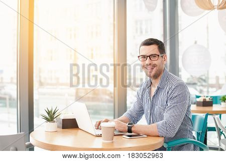 Coffee time. Handsome young man in cafe with big window. Man with cup of coffee to go. Man with glasses working with laptop, looking at camera and smiling