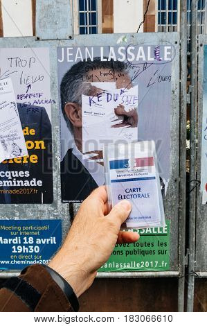 STRASBOURG FRANCE - APR 23 2017: French voter registration card held by male hand in front of official campaign poster of Jean Lassalle candidate for the 2017 French presidential elections posted outside a polling station