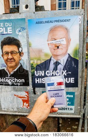 STRASBOURG FRANCE - APR 23 2017: French voter registration card held by male hand in front of official campaign poster of Francois Asselineau candidate for the 2017 French presidential elections posted outside a polling station