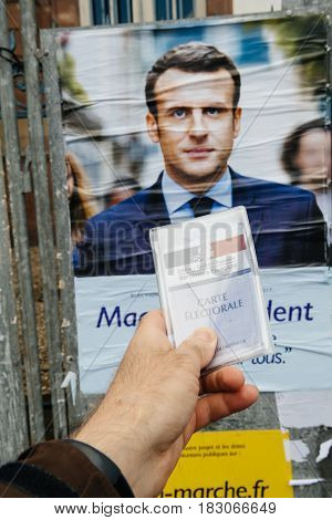 STRASBOURG FRANCE - APR 23 2017: French voter registration card held by male hand in front of official campaign poster of Emmanuel Macron candidate for the 2017 French presidential elections posted outside a polling station